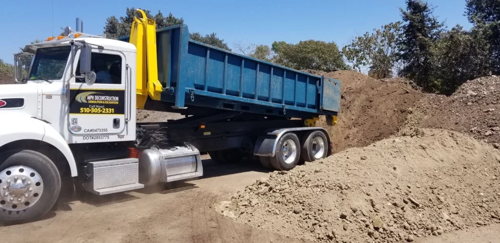 A dump truck unloading a pile of dirt at a construction site in Marin County. The truck has a blue bed and is surrounded by dirt mounds with trees in the background.