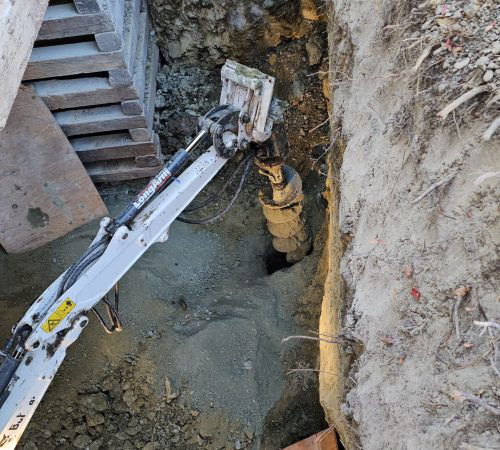 A construction site in Oakland with a small excavator digging a deep trench next to a building foundation. Various debris and pipes are visible in the trench.