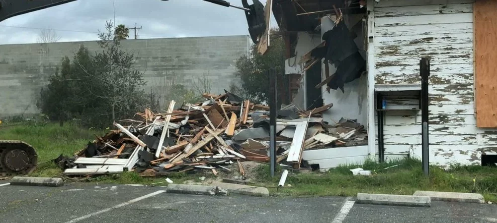 A yellow excavator demolishes a deteriorating white building with boarded windows in San Francisco, leaving a pile of wooden debris in the parking lot.