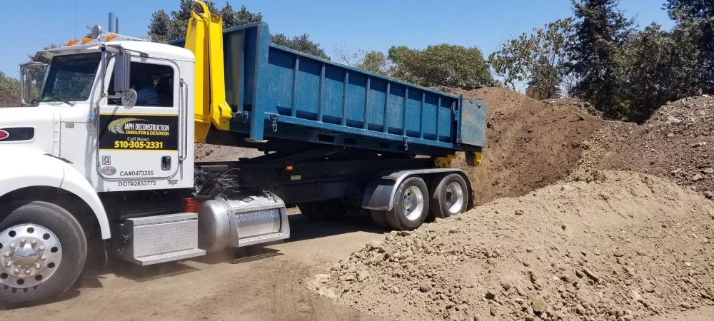 A large dump truck with a blue bed is parked on a dirt lot in Walnut Creek. The bed is raised and positioned next to a large pile of dirt on a sunny day.