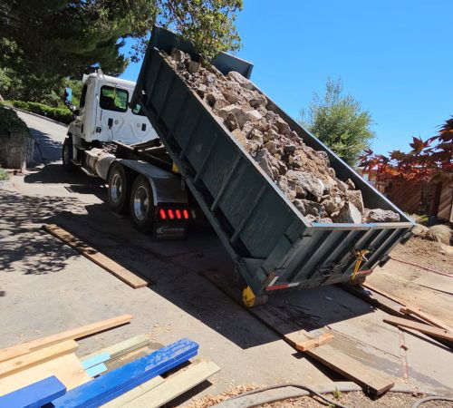 A dump truck is offloading a pile of rocks onto a sloped driveway in Walnut Creek, next to some scattered wooden planks, under a clear blue sky. Trees and bushes are visible in the background.