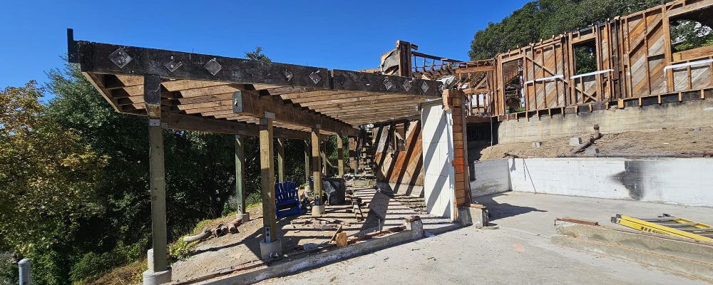 A construction site featuring a partially built structure with exposed wooden beams and supports. Trees and clear blue sky are visible in the background.