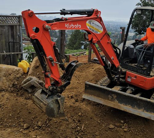 A red Kubota excavator is being used to dig into a dirt hill in Marin County. The machinery operator, wearing an orange safety vest, is inside the cab. A wooden fence and a partially cloudy sky are in the background.