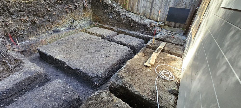 Excavated site with several large square and rectangular holes prepared for foundation work next to a building. Tools and equipment, including a wooden plank and white cord, are visible on the ground—work expertly handled by a top-rated excavation contractor.
